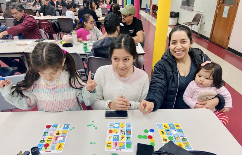 Adelante Hispanic Achievers work is pictured here. A Hispanic/Latinx mother is pictured smiling holder her baby and particpating in activities with her two additonal daughters.